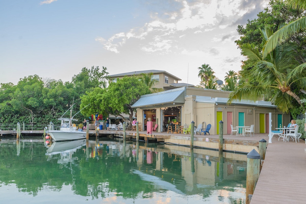 A calm waterway reflects the surrounding greenery and buildings. A wooden dock extends towards a small boat, with various seating options visible nearby. The nearby building features multiple doors and windows, indicating a welcoming atmosphere. Lush palm trees enhance the tropical setting.