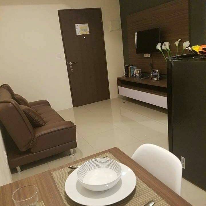 A cozy living space is presented with a brown sofa facing a flat-screen TV mounted on a wall unit. A dining table is set with a white bowl and cutlery, complementing the modern décor. The light-colored tile floor adds to the room's overall brightness.