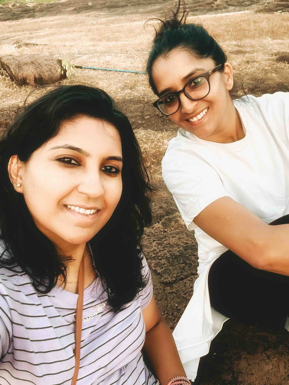 Two individuals are seated outdoors on a rocky surface, with a natural landscape visible in the background. One person is wearing a striped shirt, while the other is dressed in a white t-shirt and dark pants. The lighting appears warm, indicating a relaxed atmosphere.