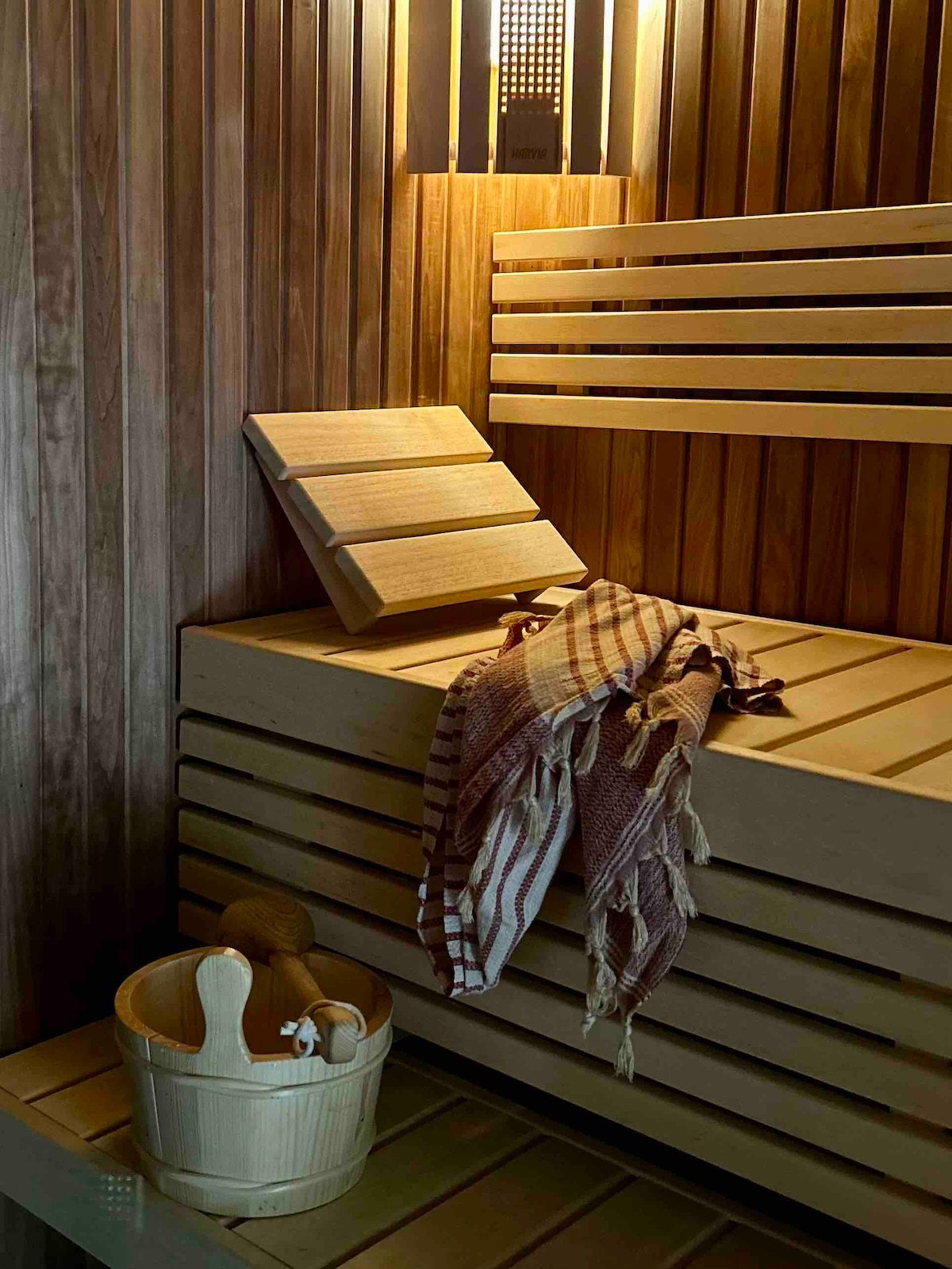 An intimate Finnish sauna is depicted, featuring wooden benches with a warm, natural finish. A woven blanket rests casually on one of the benches, and a wooden bucket and other sauna accessories are organized nearby. Soft lighting creates a calming atmosphere.