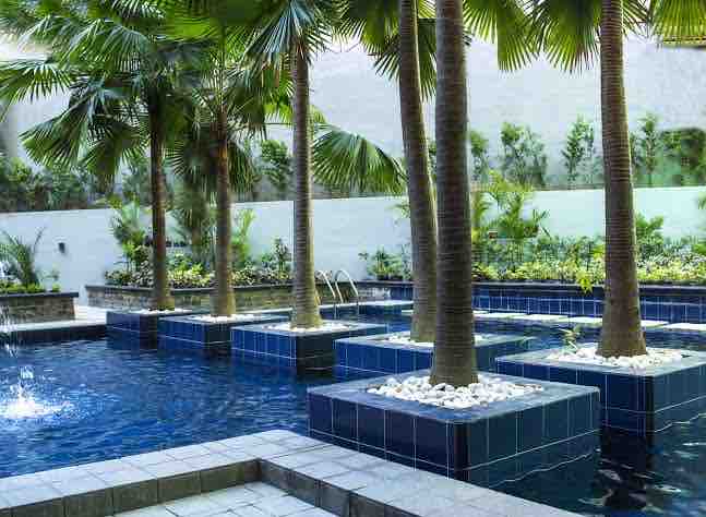 A serene pool area is surrounded by lush greenery, featuring several palm trees that provide shade. The pool is designed with dark tiles and has a few water features, enhancing the tranquil setting. Comfortable loungers are also visible in this relaxing outdoor space.