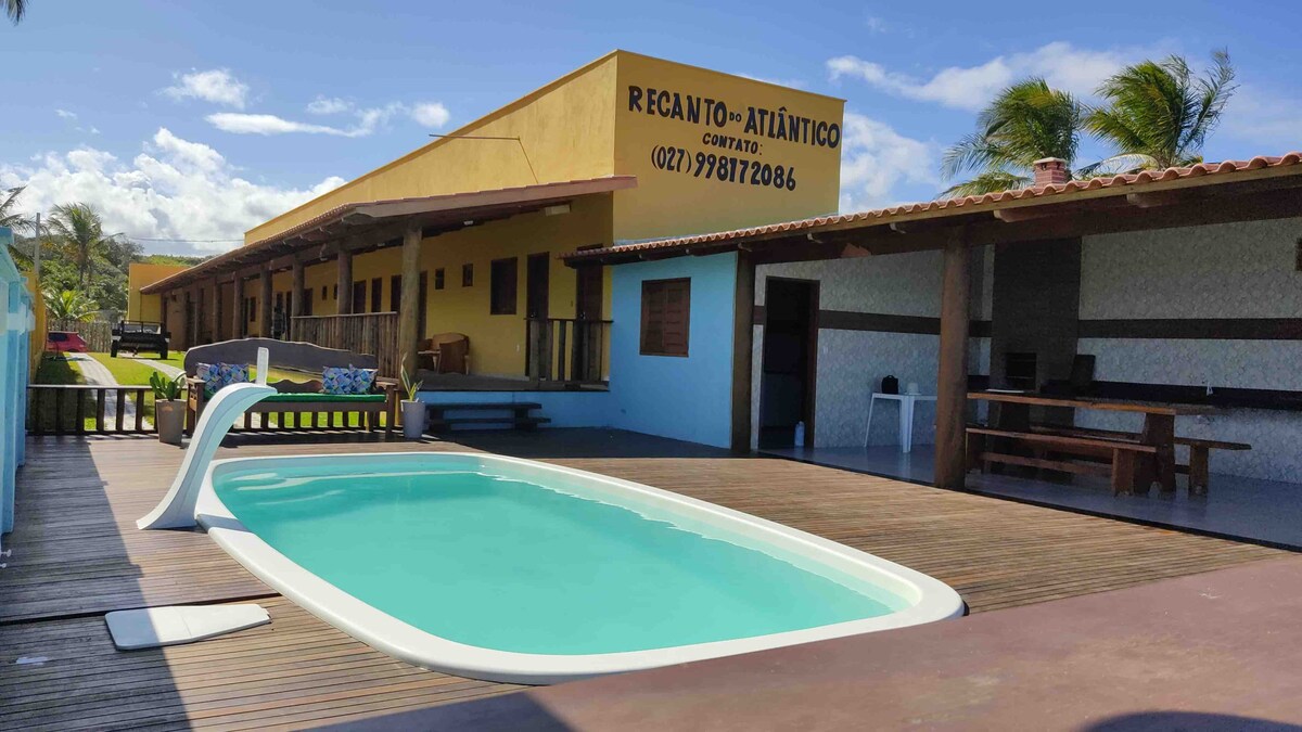 A well-maintained outdoor area features a white pool surrounded by wooden decking. The uplifting blue sky and palm trees gently sway in the background. A shaded seating area is visible, with a barbecue space designed for outdoor cooking.