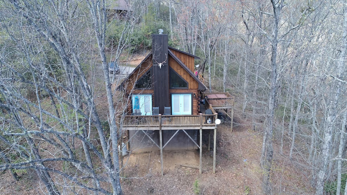 The cabin is set amidst a natural landscape, featuring a peaked roof and multiple windows that allow for natural light. A spacious deck extends from the main structure, surrounded by trees, providing a sense of privacy and connection to the outdoors.