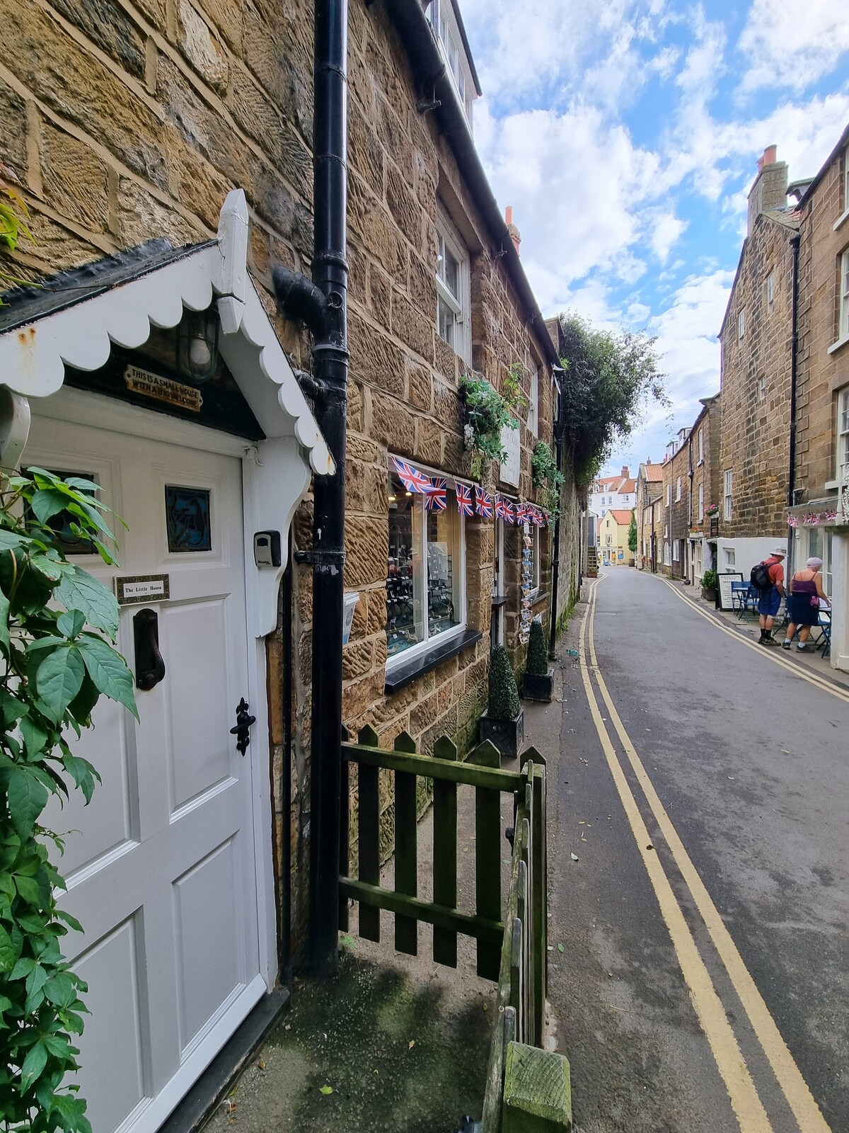 The exterior of a quaint cottage is visible, featuring a white door framed by decorative trim. Flag bunting hangs along the walls, while a narrow, cobblestone street lined with traditional stone buildings can be seen beyond. Lush greenery adds a touch of nature to the scene.