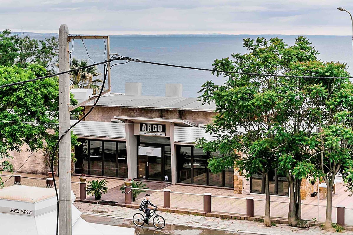 A view of a coastal establishment named 'ARGO' is presented, featuring large glass windows that reflect the serene sea. Lush green trees frame the foreground, while a cyclist is seen riding along the adjacent pathway. The calm water and cloudy sky create a tranquil atmosphere.