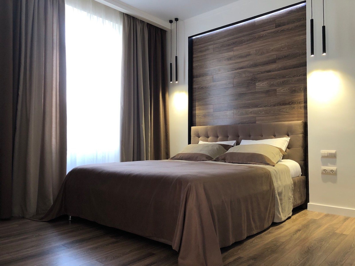 A spacious bed with a soft brown coverlet is positioned against a wood-paneled feature wall. Soft lighting from two pendant lights accentuates the neutral tones in the room. Floor-to-ceiling curtains frame a large window, allowing natural light to enter.
