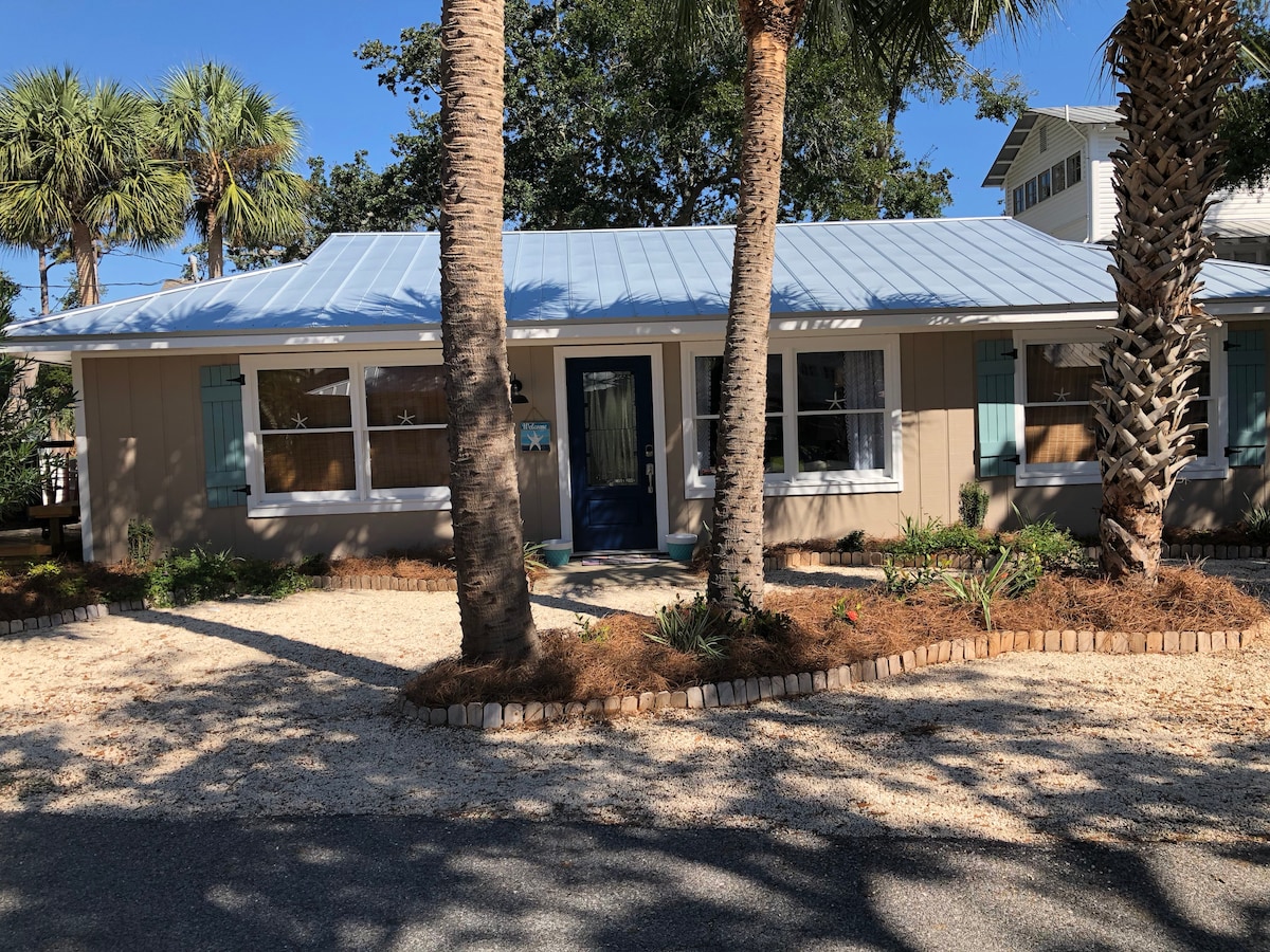The cottage features a charming exterior with light-colored siding and blue shutters. It is flanked by palm trees and surrounded by a landscaped yard with natural mulch. A welcoming blue front door is positioned at the center, inviting guests into the home.