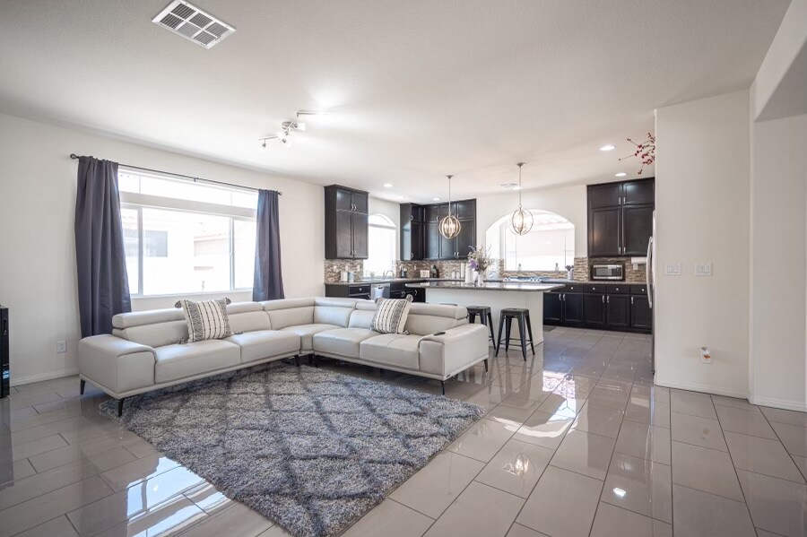 A modern living area showcases a spacious sectional sofa arranged on a soft area rug. Sleek cabinetry is visible in the open kitchen, complemented by stylish pendant lighting. Natural light floods the space through large windows, enhancing the contemporary design.