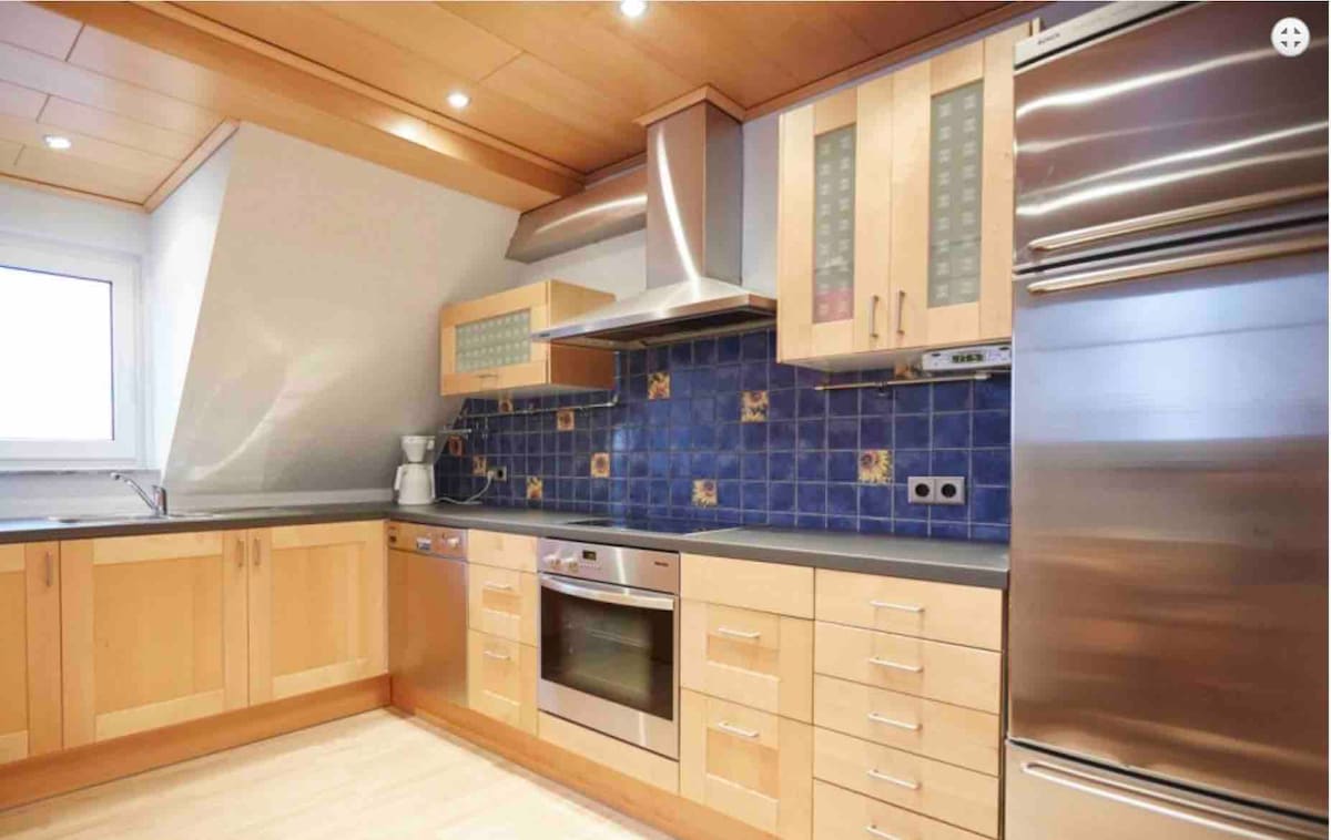 A modern kitchen features wooden cabinetry and stainless steel appliances, including an oven and refrigerator. A tiled backsplash with blue tones adds color, and ample countertop space is available for meal preparation. Adequate lighting brightens the area, highlighting its functional design.