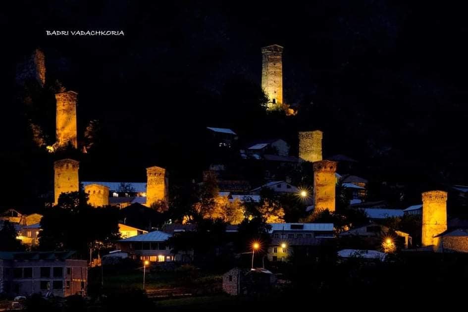 Illuminated Svan towers rise against the night sky, showcasing a historical village nestled in the valley. The warm glow of lights highlights the stone structures, while the surrounding trees and low buildings contribute to the serene atmosphere of the location.