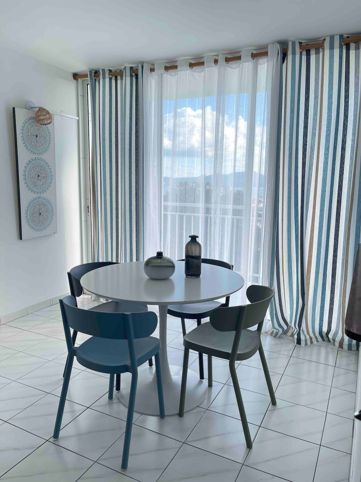A dining area is presented with a round white table surrounded by four chairs in varying shades of blue and gray. Natural light filters through sheer striped curtains, illuminating the space and providing a view of the outdoors.