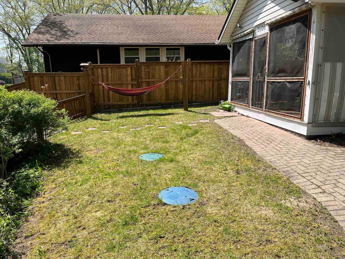 A well-maintained backyard features a grassy area with a brick path leading towards a screened-in porch. A hammock hangs between two posts, inviting relaxation. Two blue circular objects are placed on the lawn, and a wooden fence provides privacy to the space.
