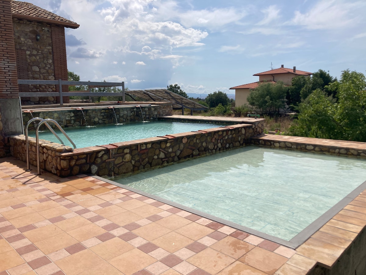 Two inviting pools are situated on a stone deck, surrounded by lush greenery. One pool features a gradual entrance with steps, while the other is slightly raised, creating a layered effect. The scenic landscape is visible in the background, enhancing the tranquil setting.