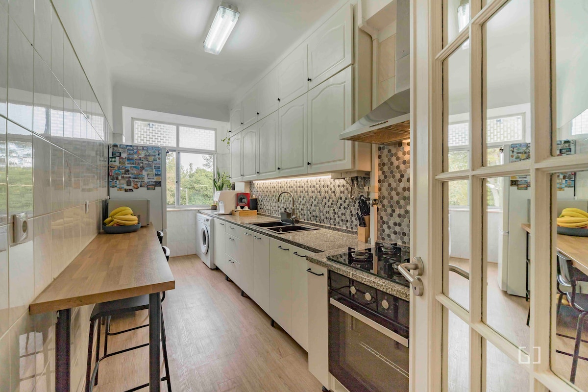 The kitchen features a clean, modern design with white cabinetry and a patterned backsplash. A countertop extends along one side, complemented by several bar stools. Natural light enters through the window, illuminating the space. Appliances include a washing machine and a stove.