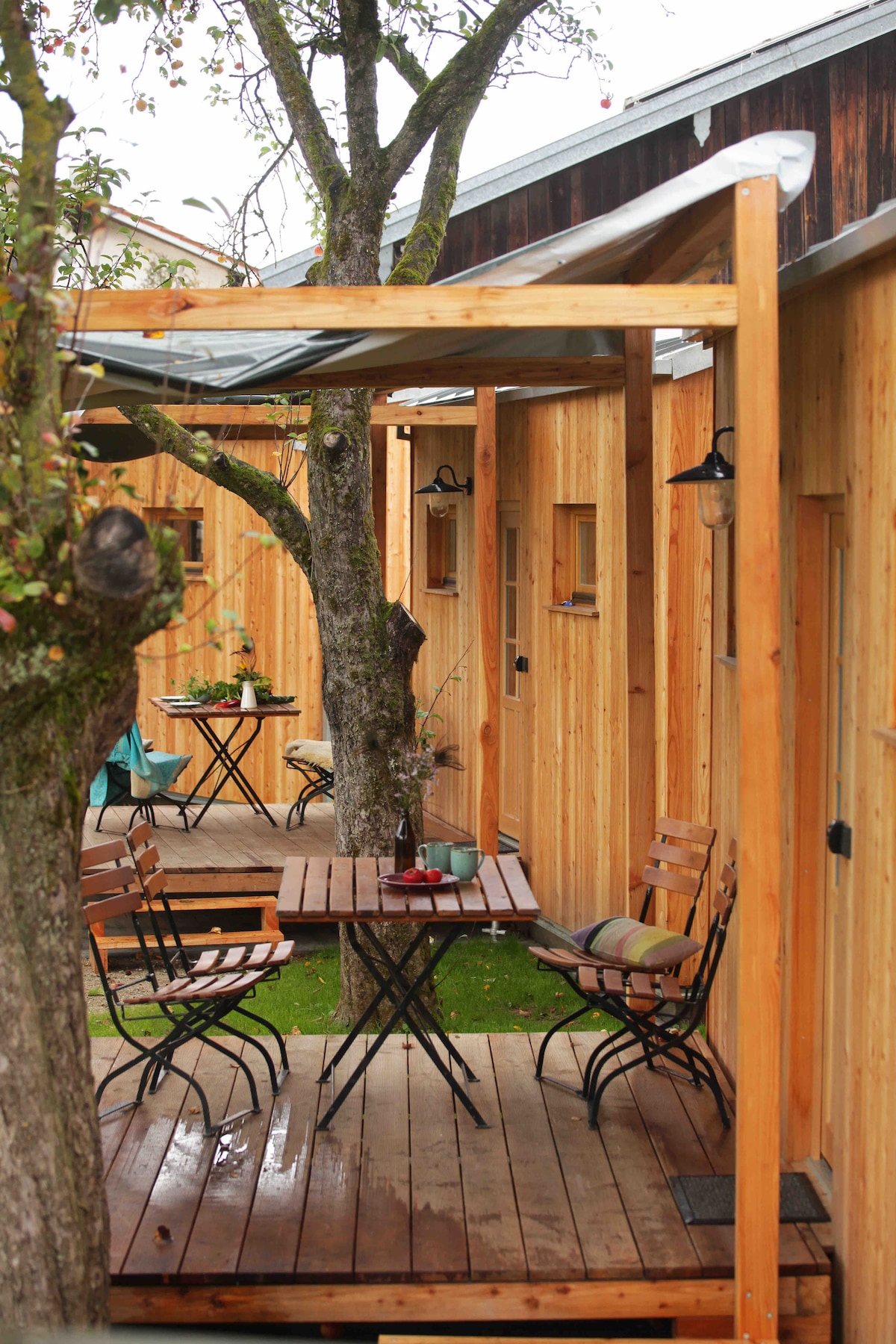 A wooden terrace is captured, featuring a table surrounded by several chairs. The area is partially sheltered, with a tree integrated into the design. Nearby, additional seating can be seen, suggesting a relaxed outdoor space ideal for socializing.