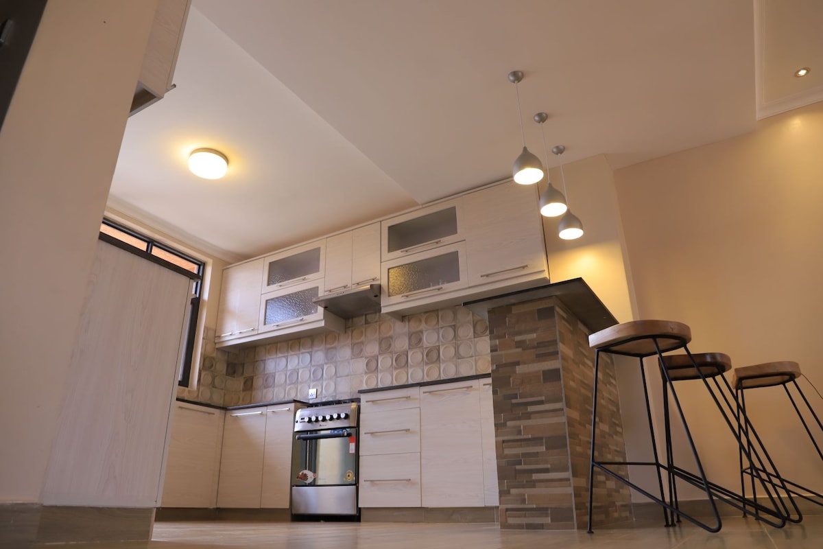 The kitchen features streamlined cabinetry, a modern gas stove, and a spacious countertop. Three tall bar stools are positioned at the kitchen island, and a window allows natural light to illuminate the area. Warm light fixtures provide additional lighting, enhancing the functionality of the space.
