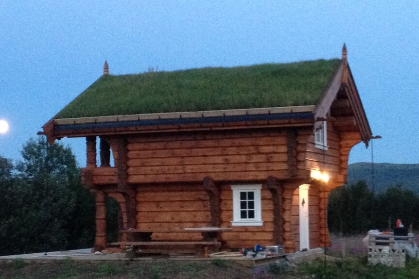 A wooden cabin features a grass-covered roof, blending with the natural surroundings. The structure showcases intricate log construction, with decorative elements visible at the corners. A small white window and a door provide access, while a picnic table is situated in front of the cabin.