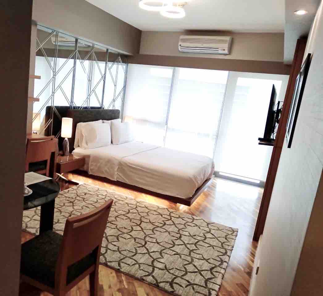 A well-appointed studio apartment features a spacious bed with white bedding and four pillows. A modern wooden table with chairs is visible in the corner, complemented by a soft area rug. Large windows allow natural light to illuminate the room, and a flat-screen TV is mounted on the wall.