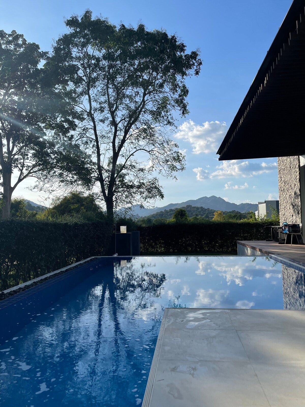 A reflective swimming pool is framed by lush greenery and trees, with mountain views visible in the background. The clear blue sky is dotted with clouds, casting soft light onto the water's surface, contributing to a serene outdoor atmosphere.