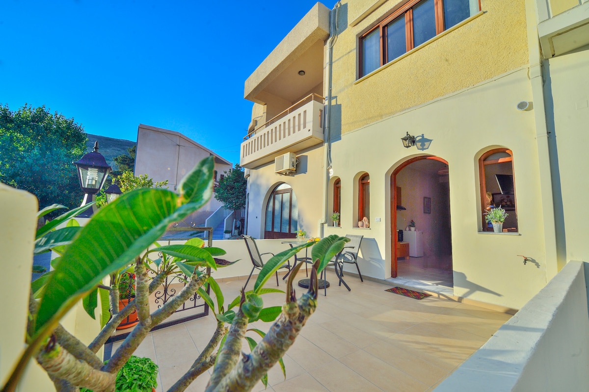 The exterior of Theofilos Studio features a welcoming terrace adorned with potted plants and a seating arrangement of chairs and a table. A pathway leads to the entrance, with large windows allowing natural light into the space. The building exhibits a blend of modern and traditional architecture.