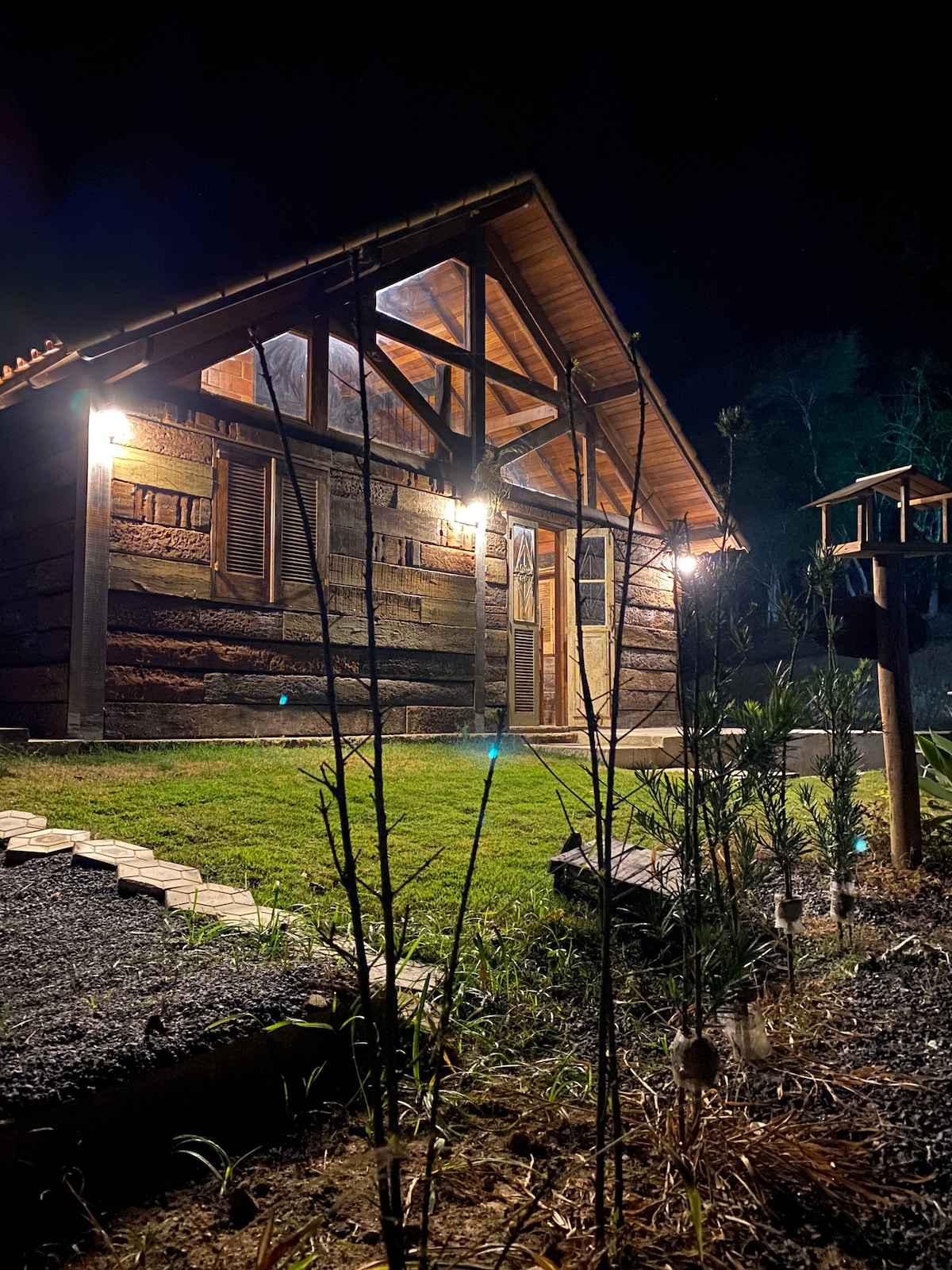 The charming cabin is illuminated softly at night, showcasing its rustic wooden exterior and distinctive architectural features. A well-maintained pathway leads from a grassy area to the entrance, with subtle lighting enhancing the natural surroundings.