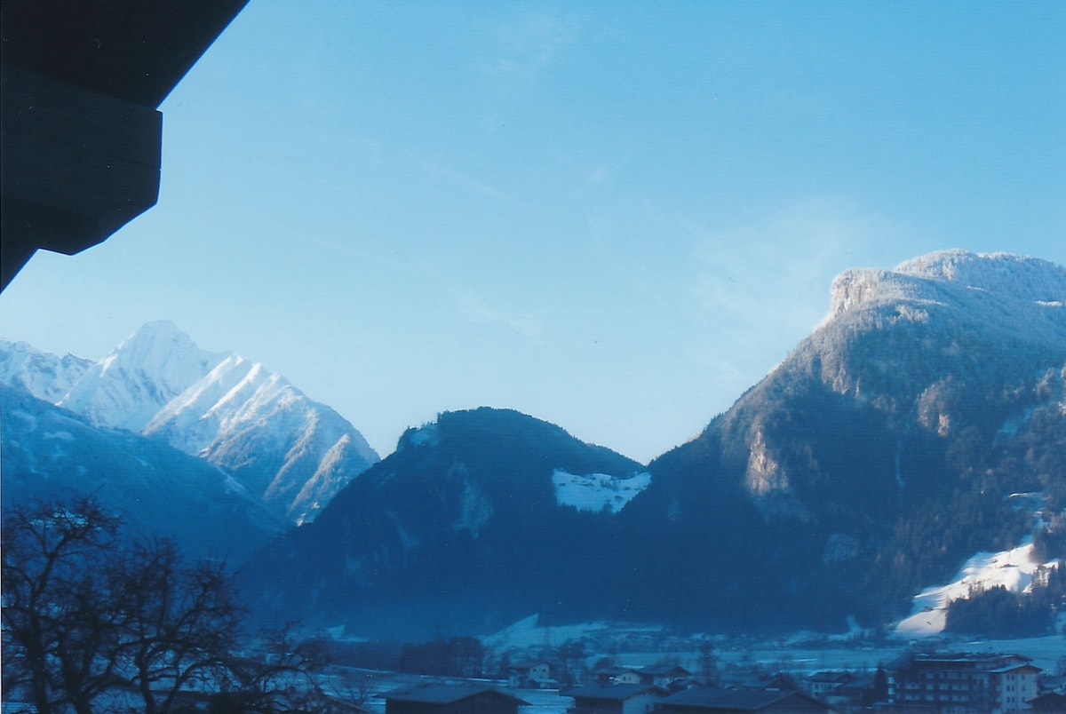 A panoramic view of the surrounding mountains is presented, showcasing snow-capped peaks under a clear blue sky. The landscape features a gentle slope and a valley with scattered houses, enhancing the natural beauty of the area.