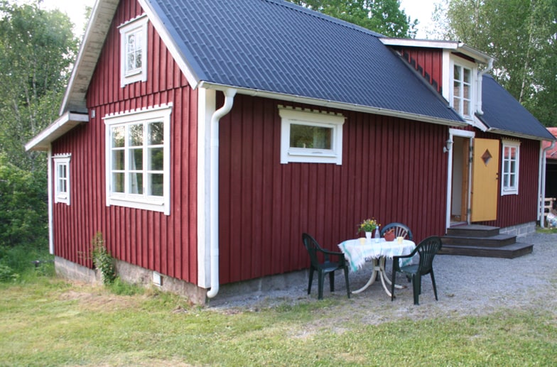 A red wooden cottage is shown with a simple, well-maintained exterior surrounded by greenery. The entrance features a warm yellow door. In front, a small gravel area is set with a table and four chairs, creating an inviting space for relaxation.
