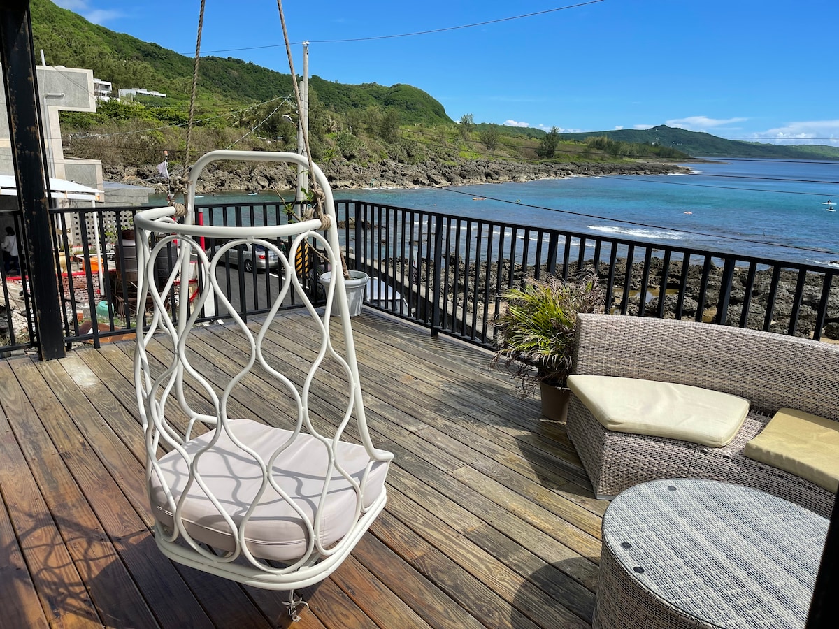 A relaxing outdoor space features a swing chair with soft cushions, positioned on a wooden deck overlooking the ocean. Surrounding the area is a comfortable seating arrangement with a round table and lush greenery, while gentle waves can be seen in the background.
