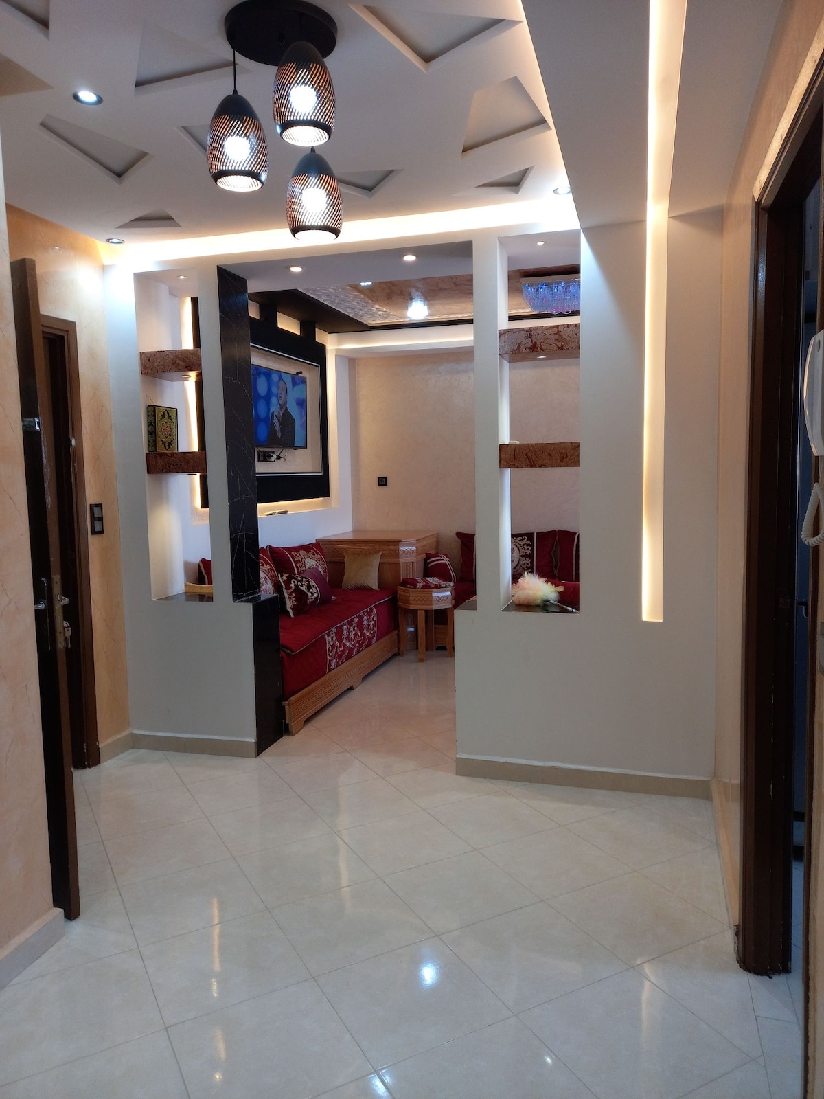 A well-lit hallway leads to a welcoming seating area. Decorative lighting fixtures hang from the ceiling, illuminating the space. The walls display modern design elements, and a pair of comfortable sofas are arranged with accent pillows. Neutral tiles create a seamless flow throughout the area.