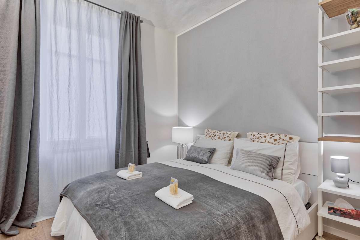 A softly lit bedroom features a spacious bed adorned with neutral bedding and two neatly arranged towels. Light gray walls complement sheer curtains, allowing natural light to brighten the space. Nightstands with lamps add a touch of warmth, enhancing the calming atmosphere.