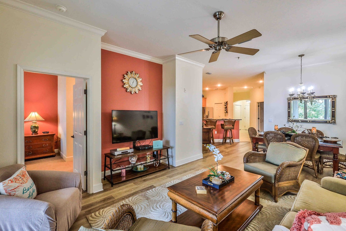 The living area features a large flat-screen TV mounted on the wall, with a comfortable seating arrangement that includes a sofa and chairs. A dining area is visible in the background, along with decorative elements like a wall clock and table lamp. Natural light fills the space.