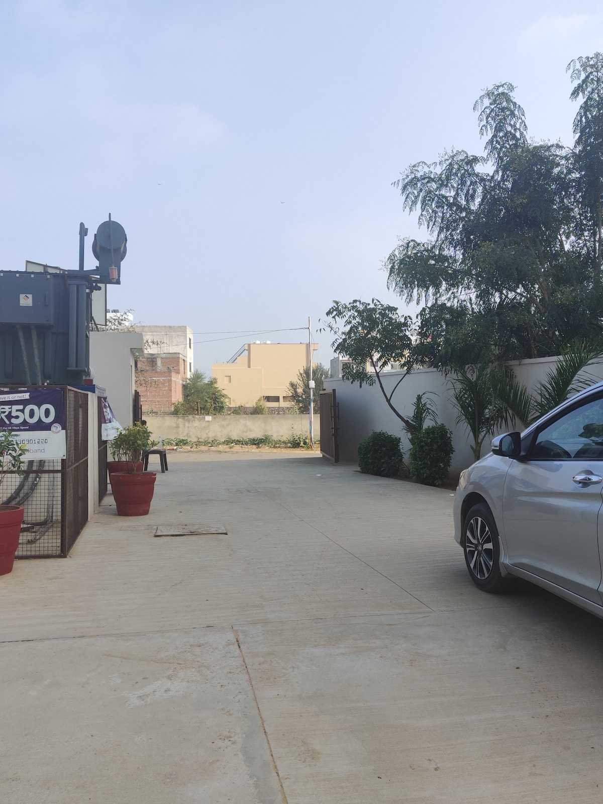 The entrance of the property is displayed, featuring a paved driveway leading to a gate. Potted plants add greenery to the setting. A parked silver car is visible, with a clear view of the surrounding area and an open sky.