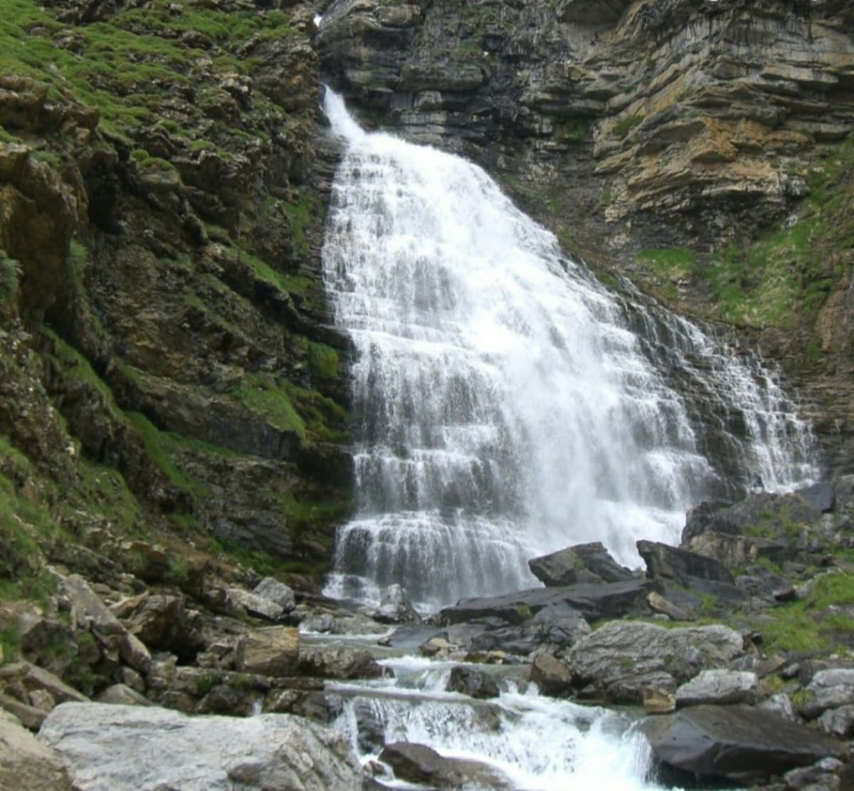A cascading waterfall flows down a rocky cliffside, surrounded by lush greenery. Water splashes against the stones below, creating a serene and natural soundscape. The scene reflects a blend of rugged terrain and smooth water, inviting a sense of tranquility.