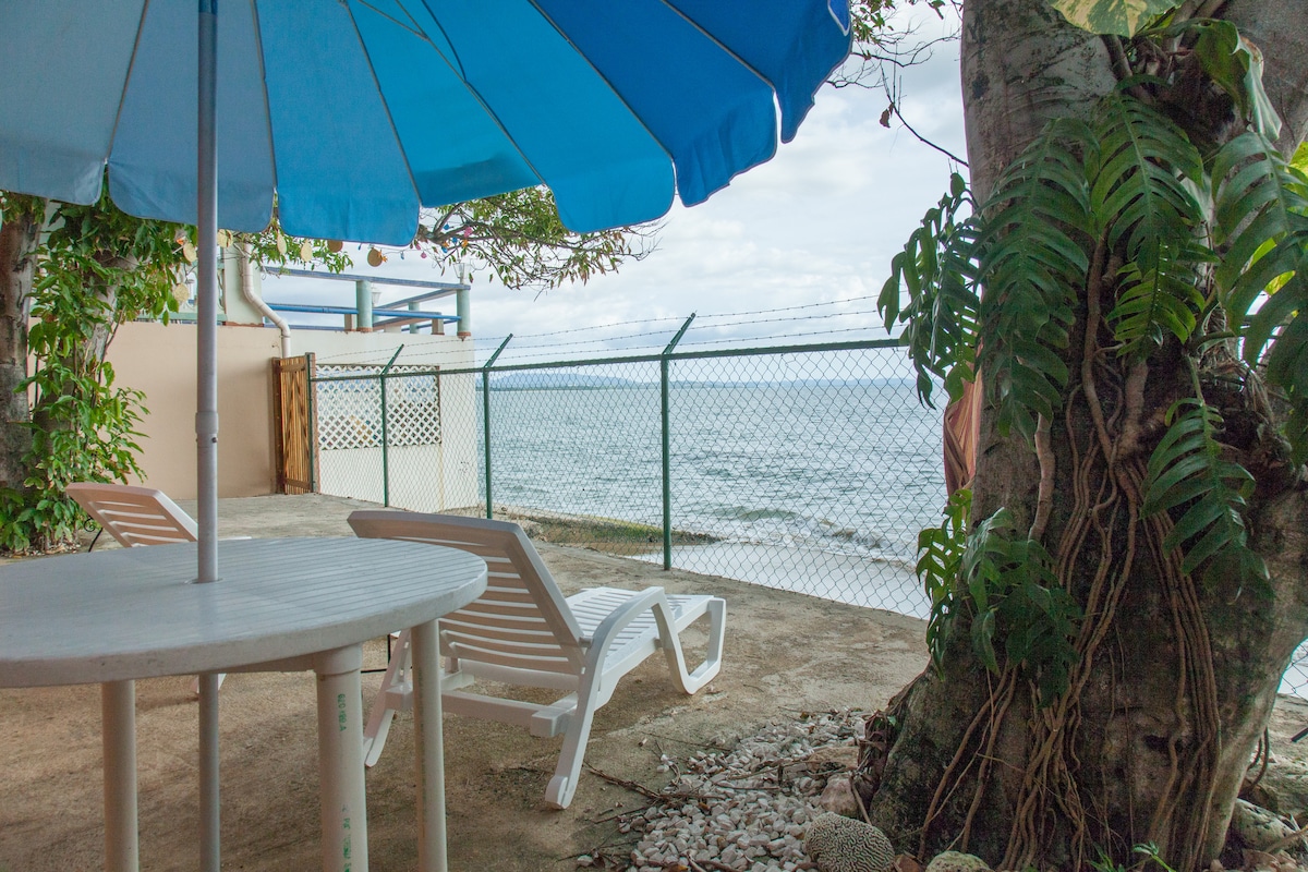 An outdoor area features a large blue umbrella providing shade over a round table and two lounge chairs. Beyond the fenced space, a view of the ocean is visible, with gentle waves lapping at the shore.