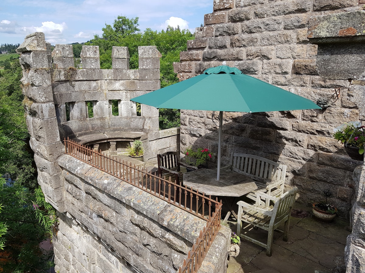 A stone terrace features a round table surrounded by wooden chairs and a built-in stone seating area. A large green umbrella provides shade, while potted plants add color. The picturesque rural landscape is visible in the background, enhancing the serene outdoor setting.