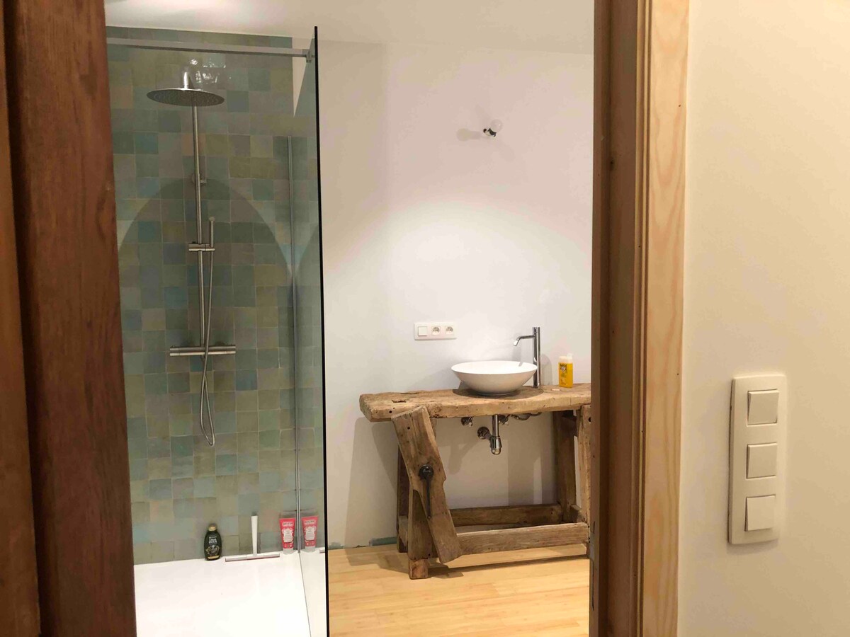 A modern bathroom is highlighted by a glass walk-in shower and a freestanding sink mounted on a rustic wooden table. Natural light illuminates the space, showcasing earthy tones in the tiled wall and light wooden flooring.