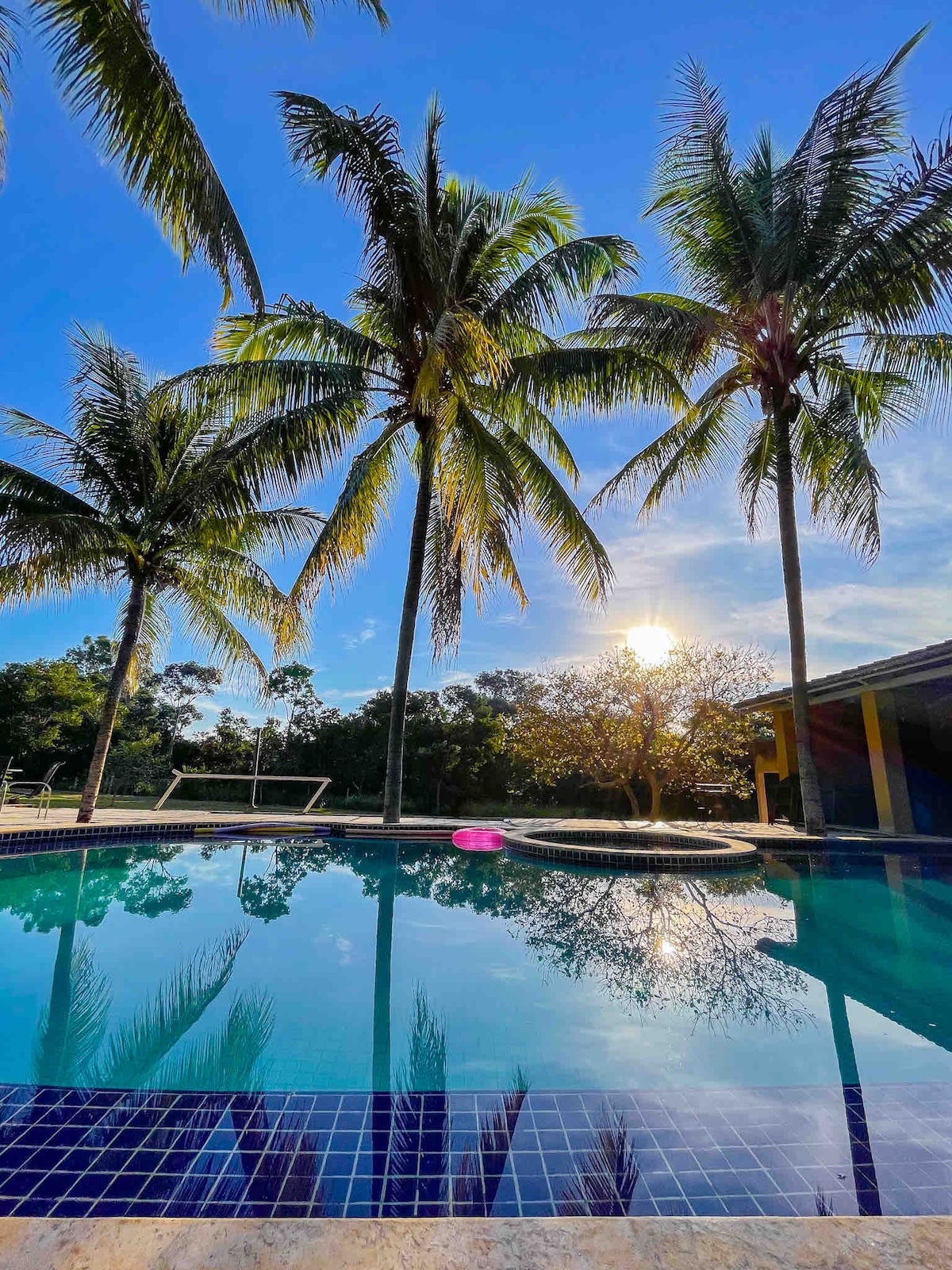 A serene outdoor pool reflects clear blue skies and palm trees, set against a backdrop of lush greenery. The sun can be seen rising, casting a warm light over the tranquil scene, creating a peaceful environment for relaxation.