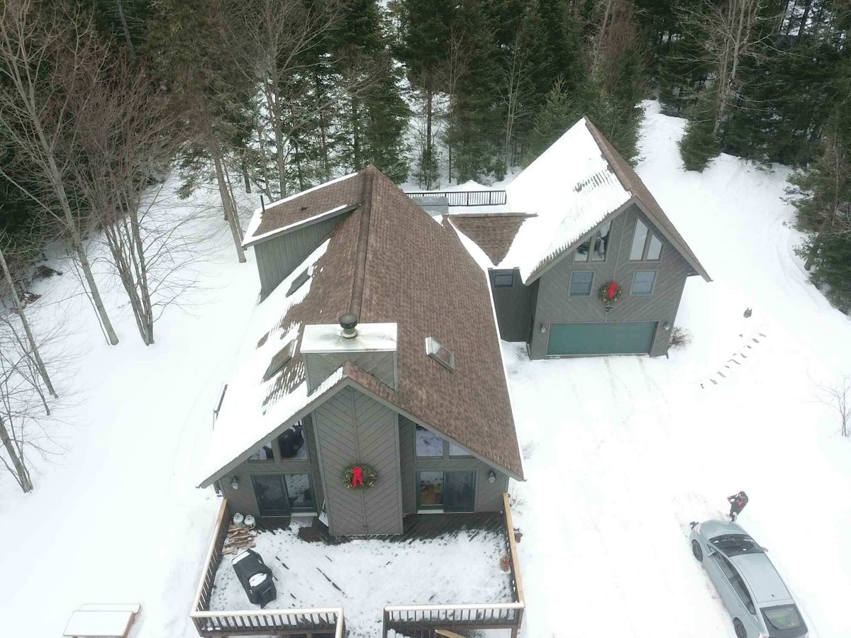 An aerial view of the home is presented, showcasing a spacious roof and multiple gables. Snow covers the surrounding ground and a deck area. The green forest backdrop adds a natural element, while festive wreaths adorn the front of the house, creating a seasonal touch.