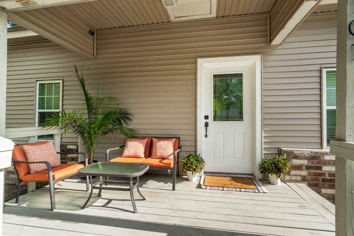 A welcoming front porch features a small table surrounded by two orange cushioned chairs and a matching loveseat. A potted plant adds a touch of greenery next to the entrance, while a decorative welcome mat lies in front of the door.