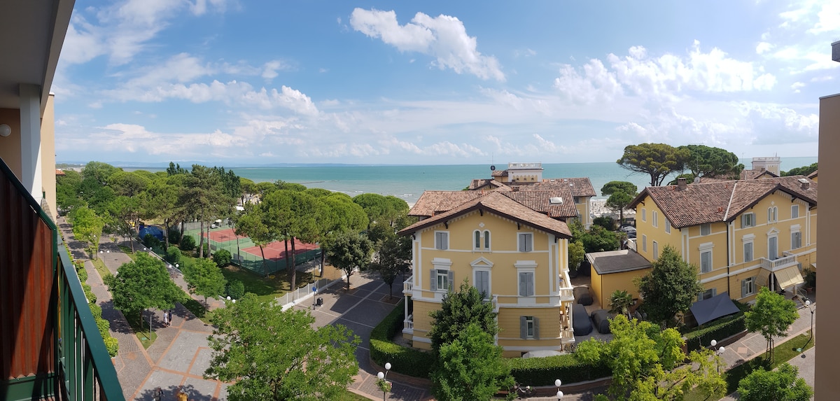 A panoramic view captures the nearby picturesque landscape, featuring charming yellow buildings and lush greenery. The serene sea stretches into the horizon, with a clear sky overhead. Tennis courts are visible amidst the trees, adding to the inviting outdoor ambiance.