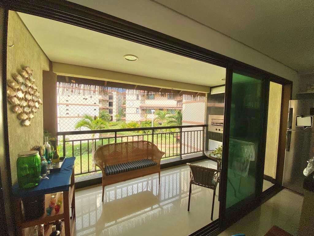A terrace area is visible, featuring a wooden bench and a small table. A substantial metal grill is positioned in the background. Lush greenery can be seen beyond the railing, and glass doors offer an open view to the outdoors.