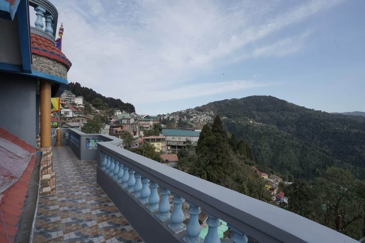 A spacious balcony is shown, offering panoramic views of the mountainous landscape in the distance. The tiled floor features a decorative pattern, and a blue railing frames the view. Green hills and structures can be seen far below, under a partly cloudy sky.