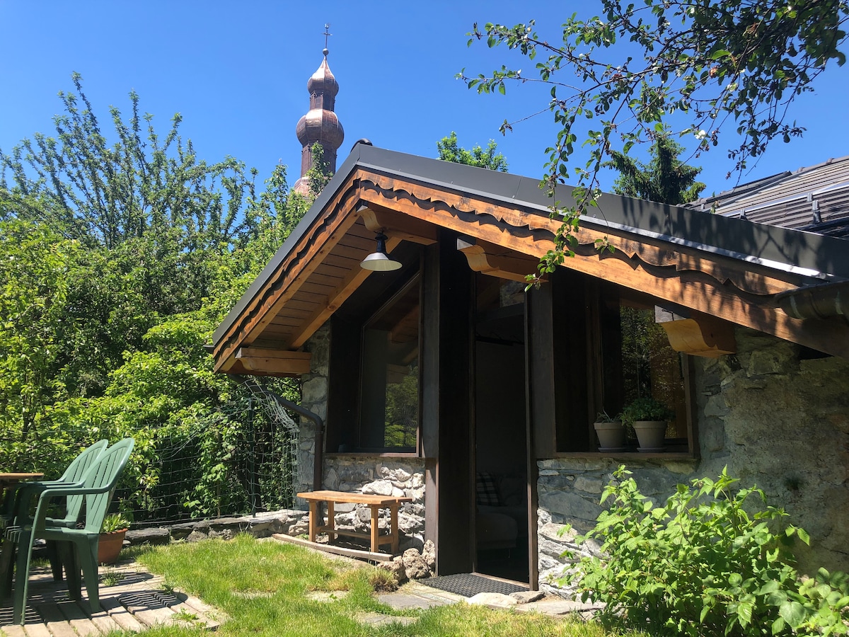 The exterior of the chalet is shown, featuring a wooden roof and stone walls. A sunny garden area is visible with green chairs and a wooden table. Lush greenery surrounds the space, adding to the tranquil atmosphere. A distinctive church tower can be seen in the background.