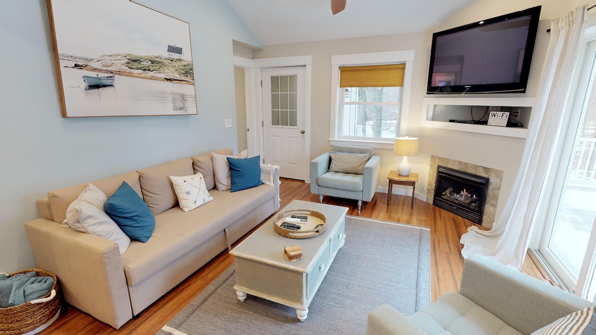 A cozy living area features a light-colored sofa with decorative pillows, paired with a comfortable armchair. A flat-screen TV is mounted above a gas fireplace, while natural light illuminates the space through a nearby window. A round coffee table sits in the center, enhancing the inviting atmosphere.