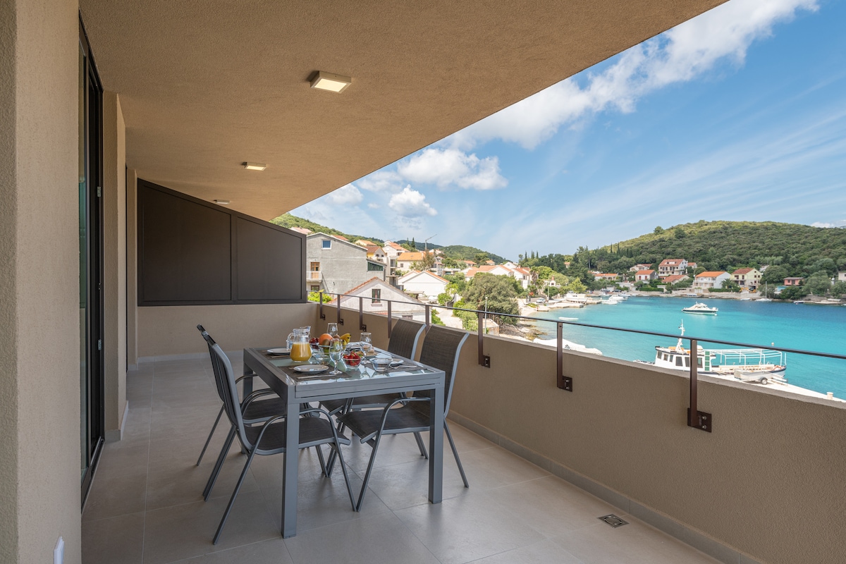 A spacious balcony dining area is presented, featuring a modern table set with an array of breakfast items. The serene view of the blue bay and distant coastal town is visible, surrounded by lush green hills under a partly cloudy sky.