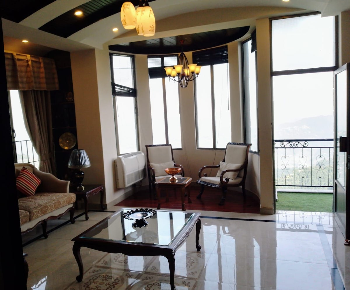 A bright living area features large windows offering views of the surroundings. Elegant furniture includes a coffee table in the center and two armchairs beside a light fixture. A cozy sofa adds to the seating options, inviting relaxation in a spacious setting.