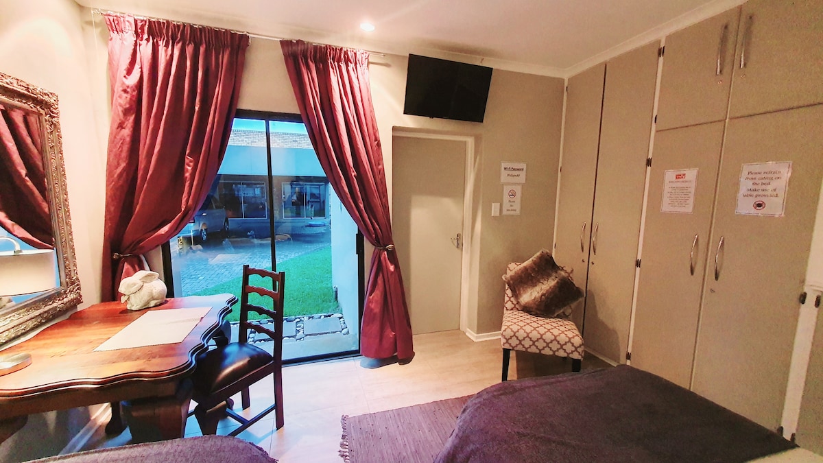 The spacious room features two single beds and a writing desk beside the window framed with rich red curtains. A television is mounted on the wall above, and a decorative chair adds to the welcoming atmosphere. A closet is positioned against the wall, and the outdoor view is visible through the sliding door.