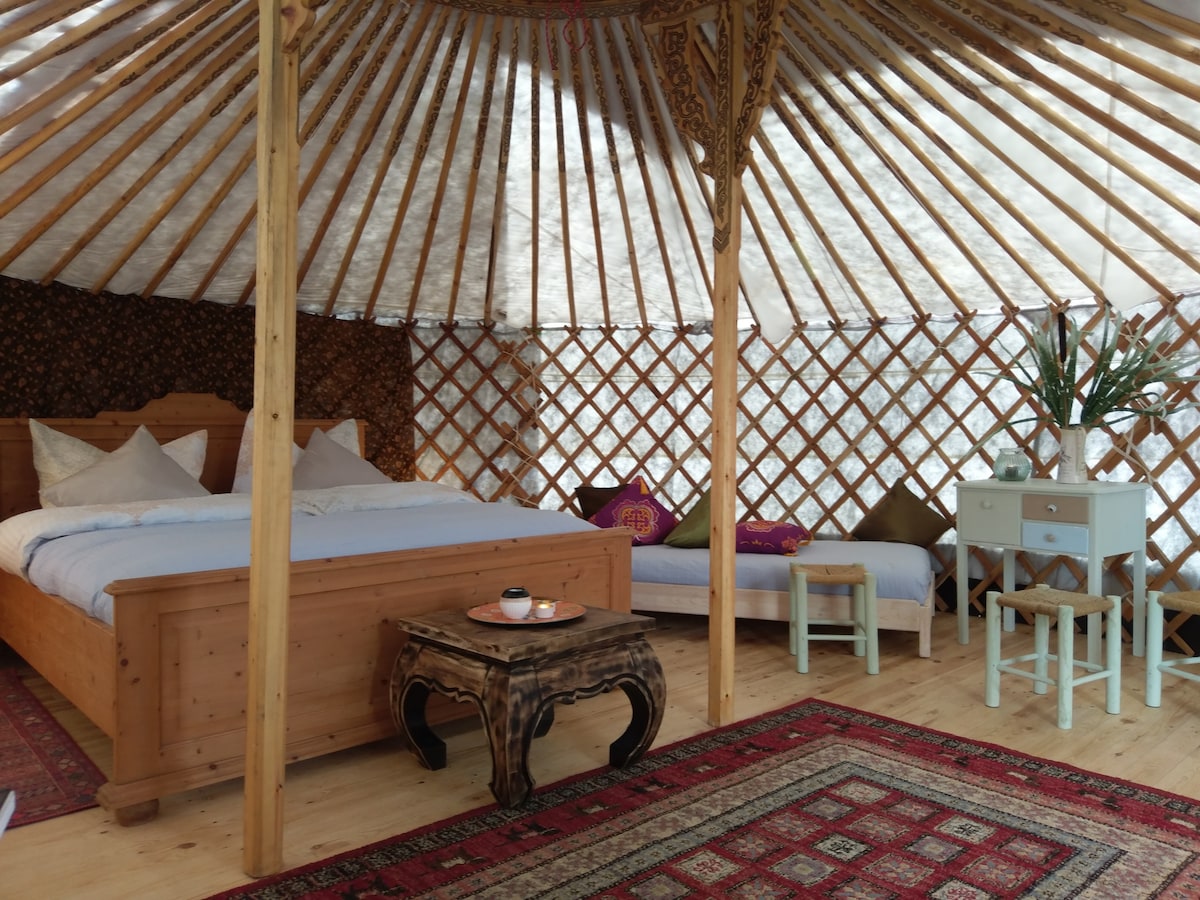 A spacious yurt interior features a wooden bed with white linens and decorative pillows. A small wooden table is positioned in the center, with a colorful rug covering the floor. A side table holds a vase with greenery, enhancing the warm ambiance.