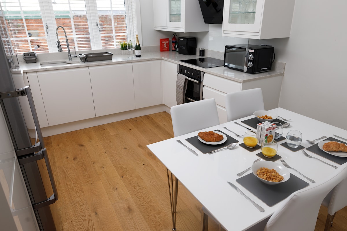 A modern kitchen is presented with sleek white cabinetry and wooden flooring. A dining table set for four features bowls of cereal, orange juice, and pastries. Large windows allow natural light to fill the space, enhancing the clean, contemporary look, while appliances are neatly arranged.