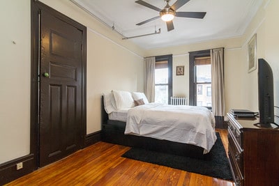 A spacious bedroom features a comfortable bed with crisp white bedding. The room is illuminated by natural light from two windows, framed with light-colored curtains. A sleek dresser and a television are positioned against the wall, while a dark rug adds warmth to the wooden floor.
